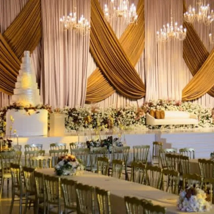 Wedding hall stage with gold and blush drapery, crystal chandeliers, white flowers and banquet tables with gold chairs