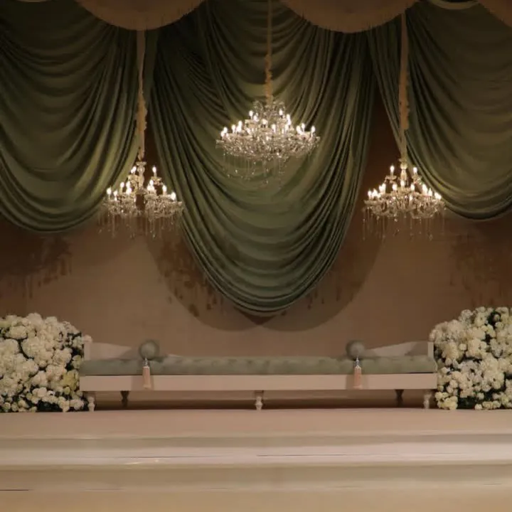 Sage green drapery with three crystal chandeliers above a tufted sofa and white flowers